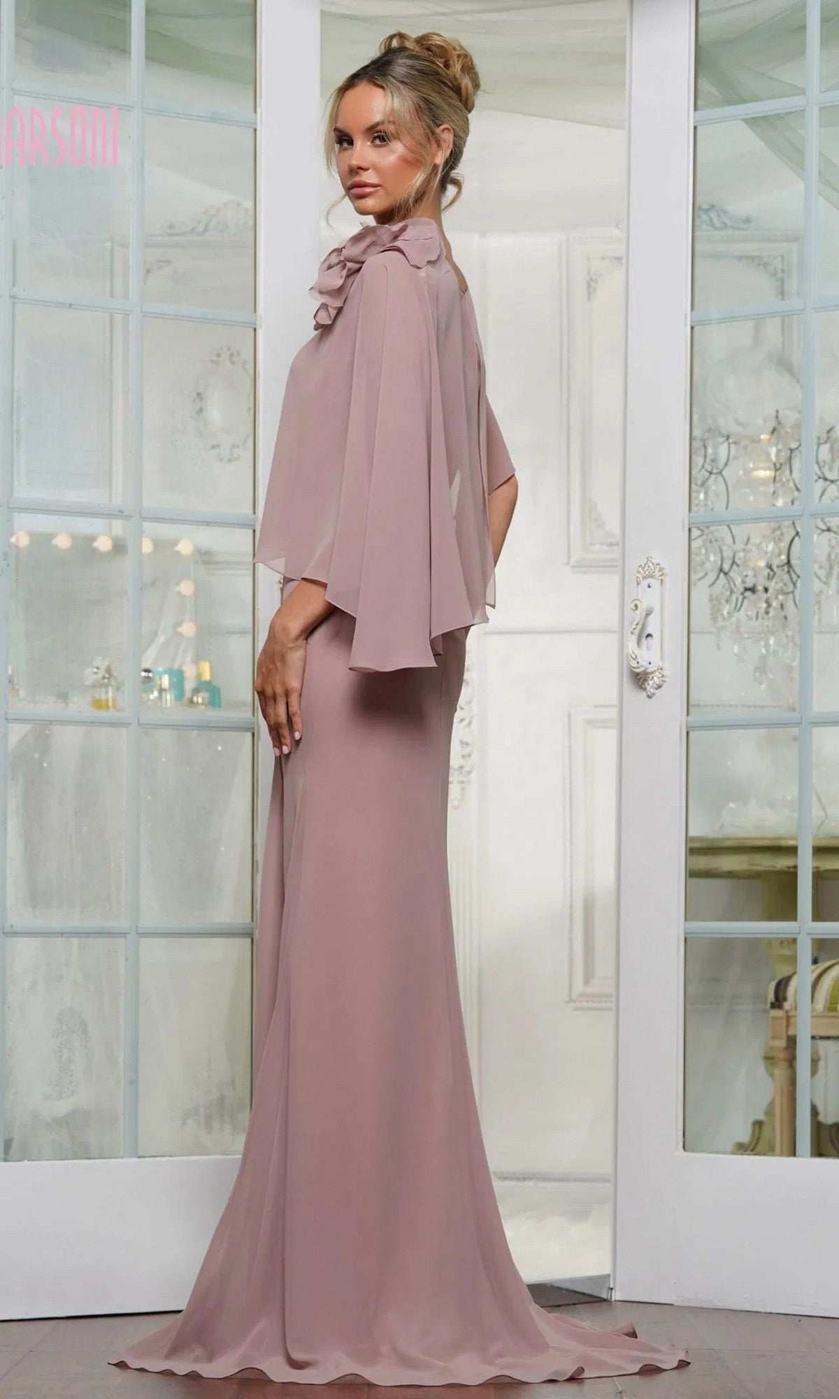 A woman in the Marsoni MV1347 long formal dress—mauve chiffon with an asymmetrical cape—stands before white double doors, glancing over her shoulder.